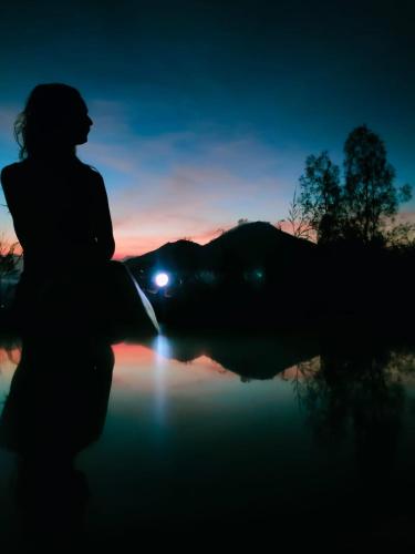 a woman sitting in front of a lake at sunset at Batur Volcano Guesthouse in Kintamani