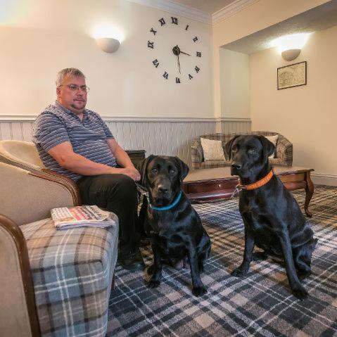a man sitting in a chair with two black dogs at Castletown Hotel in Castletown