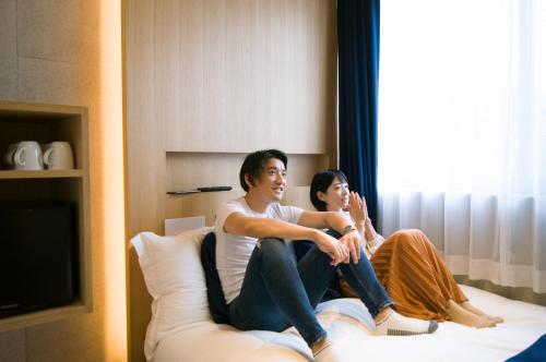 two people sitting on a bed looking out a window at Sakura Cross Hotel Shinjuku East Annex in Tokyo