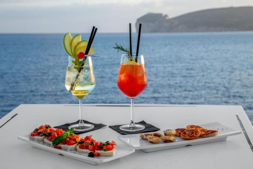 two glasses and a plate of food on a table by the water at El Faro Hotel & Spa in Porto Conte