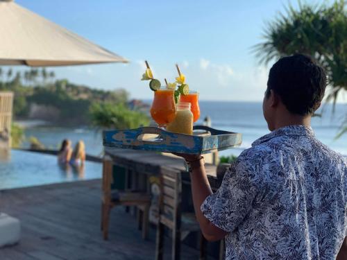 a woman holding a tray with drinks on a table near a pool at The Palms Ceningan Hotel in Nusa Lembongan