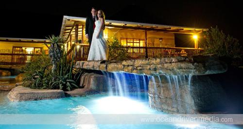 a bride and groom standing next to a waterfall at Rain Farm Game Lodge in uMhlali