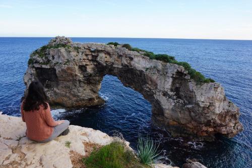 a woman sitting on a cliff looking at an arch in the ocean at Apartamentos Villa Primera in Cala Figuera