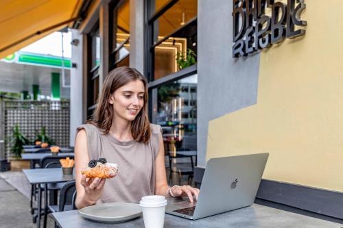 une femme assise à une table avec un ordinateur portable et une pâtisserie dans l'établissement Perouse Randwick by Sydney Lodges, à Sydney