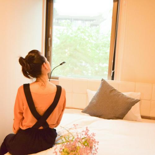 a woman sitting on a bed looking out a window at QuintessaHotel TokyoHaneda Comic&Books in Tokyo