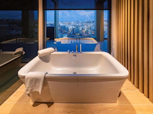 a white bath tub in a bathroom with a large window at Mercure St. Julian's Malta in St. Julianʼs