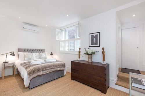 a white bedroom with a bed and a wooden dresser at 2 bed garden flat with air con by Fulham Broadway in London