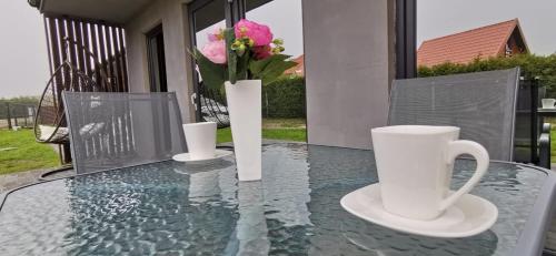 uma mesa de vidro com duas chávenas brancas e flores em Port Royal Luksusowe Domki Apartamentowe em Sarbinowo