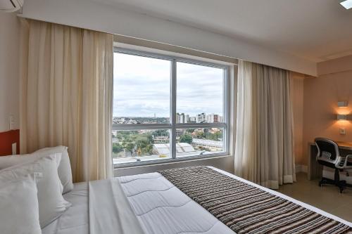 a bedroom with a large window with a bed and a desk at Ramada Hotel & Suites Campos Pelinca in Campos dos Goytacazes