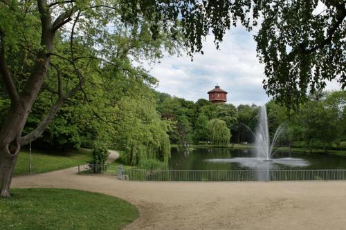 a pond with a fountain in a park with a tower at Parkhotel Altes Kaffeehaus in Wolfenbüttel