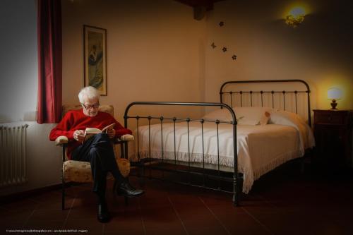 a woman sitting in a chair reading a book next to a bed at All'Antica Scuderia Del Castello in Maiano