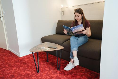 a girl sitting on a couch reading a book at Brit Hotel Essentiel Cholet in Cholet