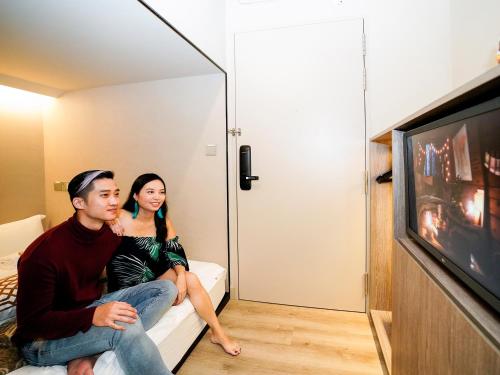 a man and woman sitting on a bed watching tv at ST Signature Jalan Besar, DAYUSE, 5 Hours, 5PM-10PM in Singapore