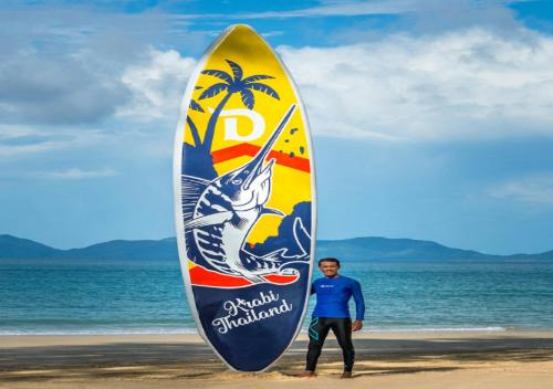 
a man standing next to a surfboard on a beach at Dusit Thani Krabi Beach Resort - SHA Extra Plus in Klong Muang Beach
