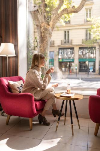 a woman sitting in a chair looking out a window at The Originals City, Hôtel Mondial, Perpignan in Perpignan