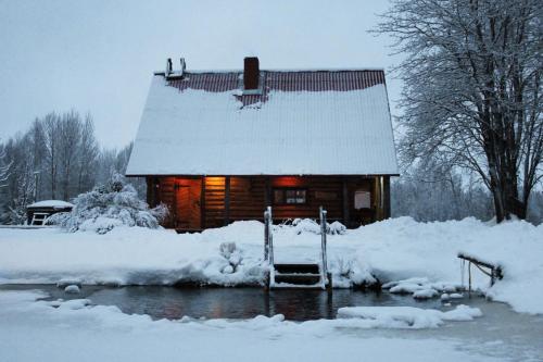 une cabane en rondins dans la neige avec les lumières allumées dans l'établissement Pirts Baudas, à Sīveci