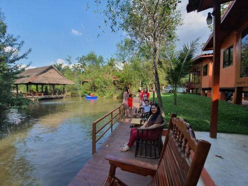 a group of people sitting on a bench next to a river at บ้านสวนในฝัน-ตลาดน้ำท่าคา in Samut Songkhram