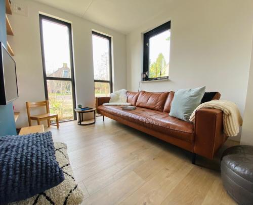 a living room with a brown leather couch and windows at Huisje Hygge - luxe bungalow met grote tuin in Ewijk