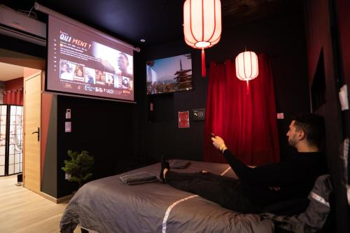 a man sitting on a bed in a room with a tv at Capsule Japon - Jacuzzi - Netflix & Ecran Cinéma - Xbox in Saint-Léger