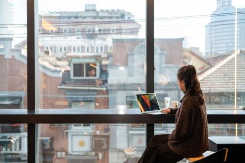 a woman sitting in front of a window with a laptop at Dayin Youth Hostel - East Nanjing Road & People's Square & The Bund -Large Social Area -Bar -Café -Gym -Free Laundry Luggage Storage -24H Front Desk -English Service in Shanghai