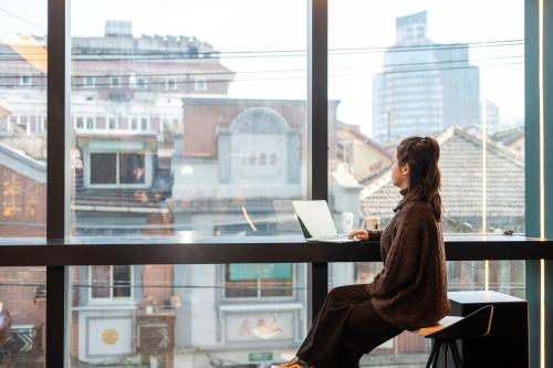 a woman sitting at a desk with a laptop in front of a window at Dayin Youth Hostel - East Nanjing Road & People's Square & The Bund -Large Social Area -Bar -Café -Gym -Free Laundry Luggage Storage -24H Front Desk -English Service in Shanghai