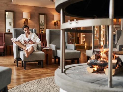 a man sitting in a chair reading a book next to a fireplace at aja Fürstenhaus am Achensee in Pertisau