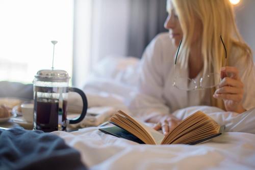 a woman laying in bed reading a book and drinking coffee at The Manor House at the Queen Victoria Hotel by NEWMARK in Cape Town