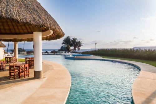 uma piscina com um guarda-sol de palha e mesas e cadeiras em Beautiful Apartment in Dream Acapulco Diamante em Acapulco