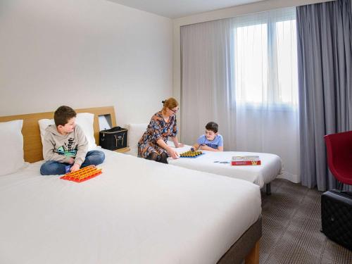 una mujer y dos niños sentados en las camas en una habitación de hotel en Novotel Bayeux, en Bayeux
