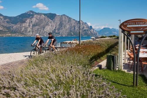 two people riding bikes on a path next to the water at Hotel Europa in Malcesine
