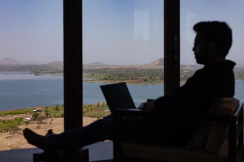 a man sitting in front of a window with a laptop at StayVista at Sunset on the Lake - Infinity Lakeview Swimming Pool with Contemporary Interiors & Lavish Amenities in Igatpuri