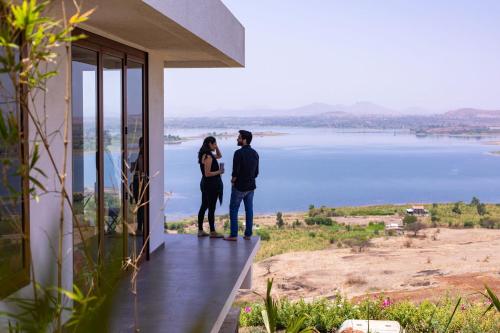 a couple standing on the balcony of a house overlooking a lake at StayVista at Sunset on the Lake - Infinity Lakeview Swimming Pool with Contemporary Interiors & Lavish Amenities in Igatpuri