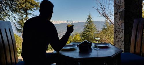 a man sitting at a table with a glass of wine at Hostie Onella - Private 3BHK Mountain Chalet with breathtaking Himalayan views in Rānīkhet