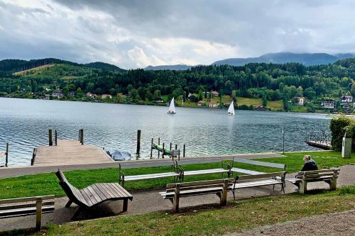 a person sitting on benches near a lake with sailboats at Maxi'Seeblick Apartments in Seeboden