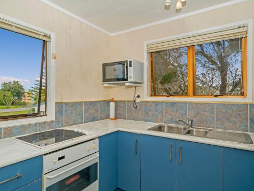 a kitchen with blue cabinets and a sink and two windows at Oyster Bach - Cooks Beach Bach in Cooks Beach