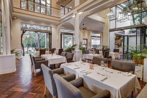 um restaurante com mesas e cadeiras brancas e janelas grandes em Palm River Hotel em Victoria Falls