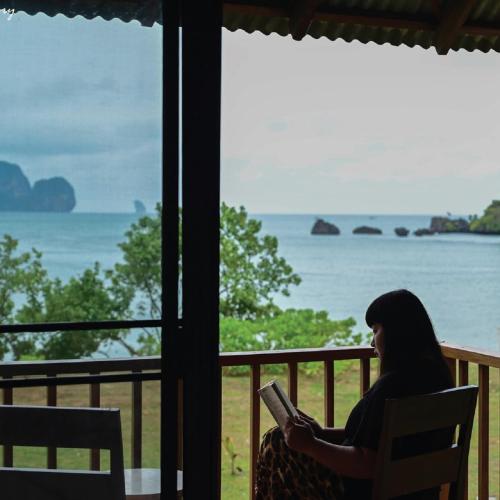 a woman sitting in a chair reading a book in front of a window at Tinidee Hideaway Tonsai Beach Krabi - SHA Extra Plus in Tonsai Beach