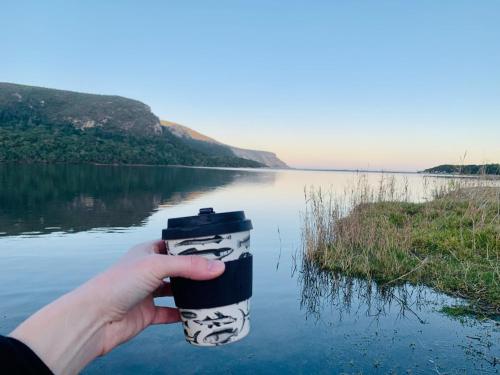 a person holding a coffee cup in front of a lake at The Wood Owl Nature's Valley in Natureʼs Valley