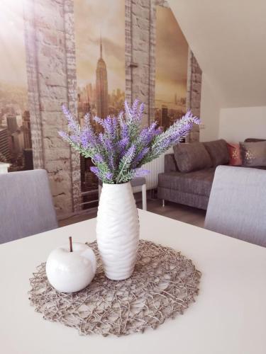 a white vase with purple flowers and an apple on a table at Ferienwohnung Lavanda in Rust