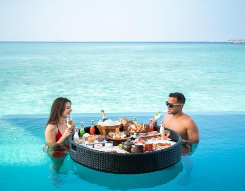 un homme et une femme assis dans l'eau avec une table de nourriture dans l'établissement Heritance Aarah-Premium All Inclusive, à Raa Atoll