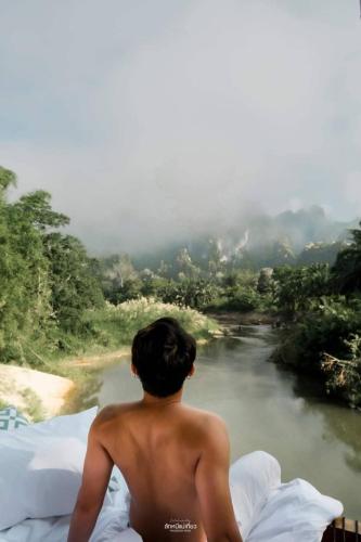 a shirtless man sitting on a bed looking at a river at Phu Siab Mhok@KhaoSok in Khao Sok