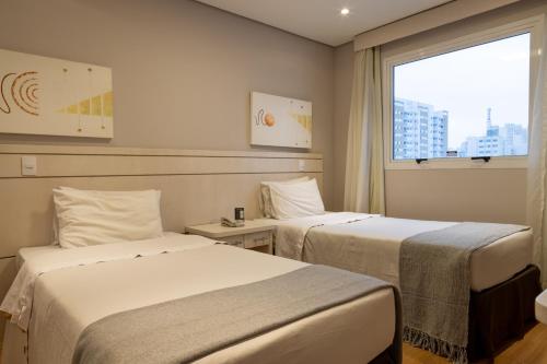 two beds in a hotel room with a window at 360 Saint Charbel - Apartamentos mobiliados in Sao Paulo