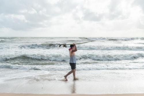 un hombre caminando por la playa con una tabla de surf en la cabeza en Kokotel Khao Lak Lighthouse - SHA Extra Plus, en Khao Lak