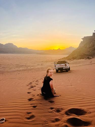 a woman sitting on the sand with a car in the desert at Shakria Bedouin Life Camp in Wadi Rum