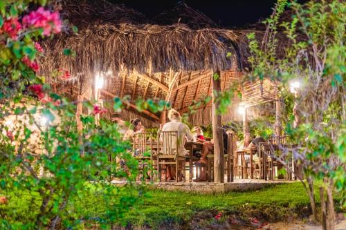 a group of people sitting at a table under a straw roof at La Cozinha Bungalow in Barra Grande