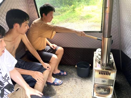 three young men sitting in front of a stove in a tent at 一棟貸切 森のお風呂とアウトドアサウナ 日光雪月花 in Nikko