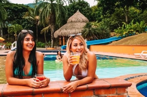 two women sitting next to a swimming pool holding drinks at Hotel Hacienda la Bonita in Amagá