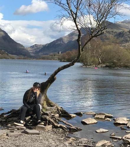 a woman sitting next to a tree on the shore of a lake at Llanberis Lodges in Llanberis