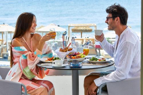 um homem e uma mulher sentados à mesa com comida e bebidas em Sea Breeze Santorini Beach Resort, Curio By Hilton em Perivolos