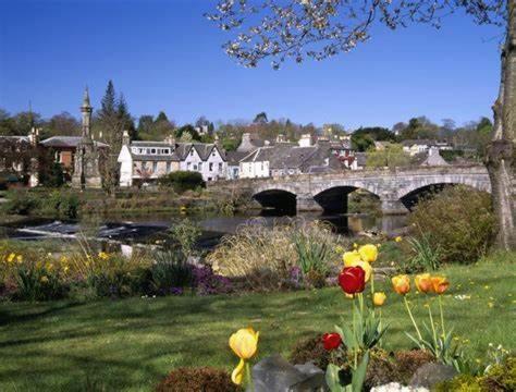 un pont sur une rivière avec des fleurs dans une cour dans l'établissement TL083 - 2 Bedrooms indoor pool Loch Views fishing Golf Riding Shooting Water Sports 15 min drive to beaches PASSES NOT INCLUDED Most Activities Will Not Be Available Out Of Season Please Check Before Booking, à Newton Stewart